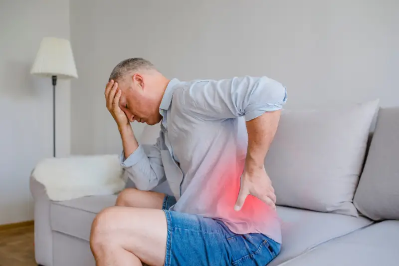 Back_pain1 A man sitting on a couch, rubbing his painful back